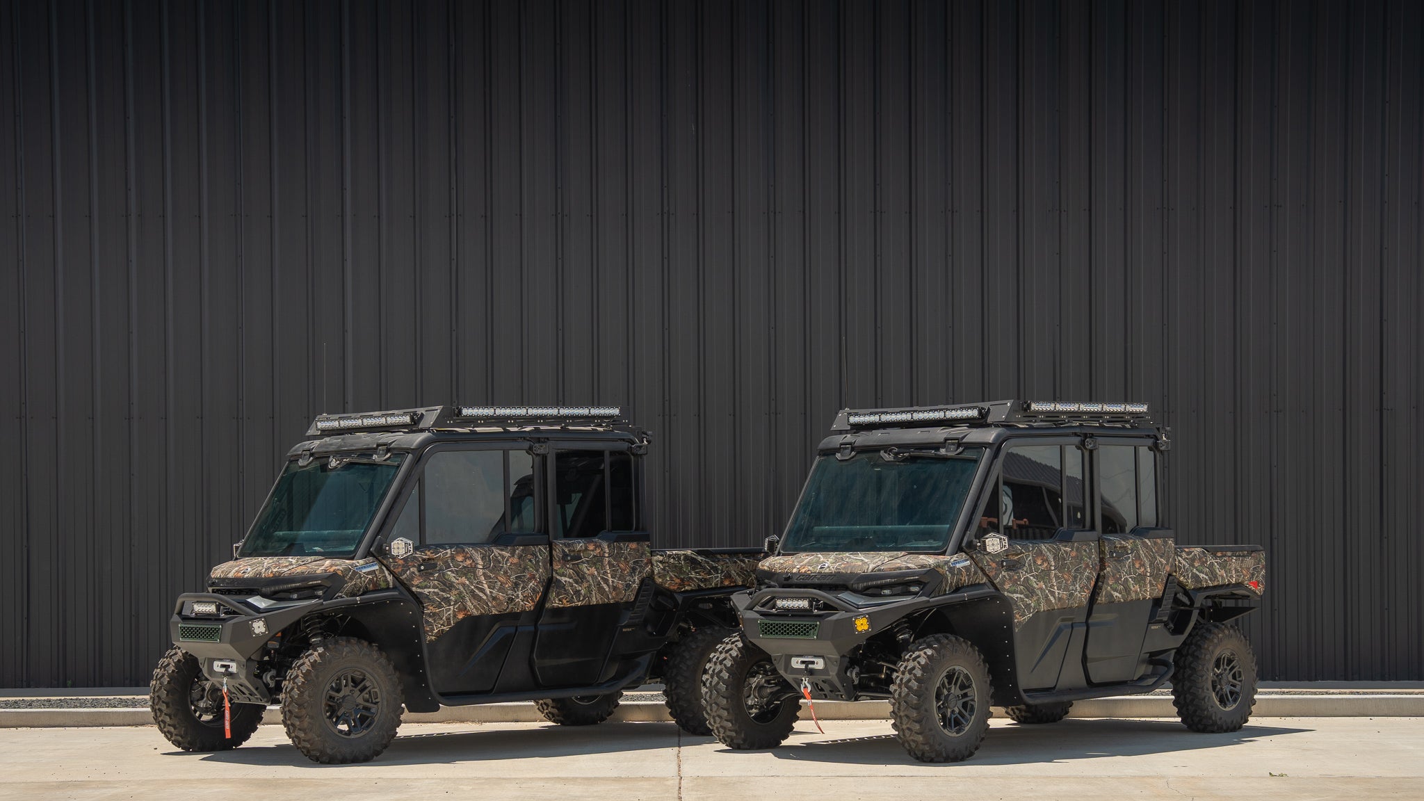 Two black off-road utility vehicles with camouflage-patterned side panels parked side by side against a dark corrugated metal wall.
